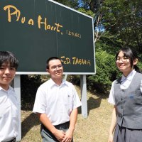学園が掲げる標語「Ｐｌｕｓ　Ｈａｕｔ」の看板の前に立つ生徒会の３人。（左から）安藤君、川名君、工藤さん